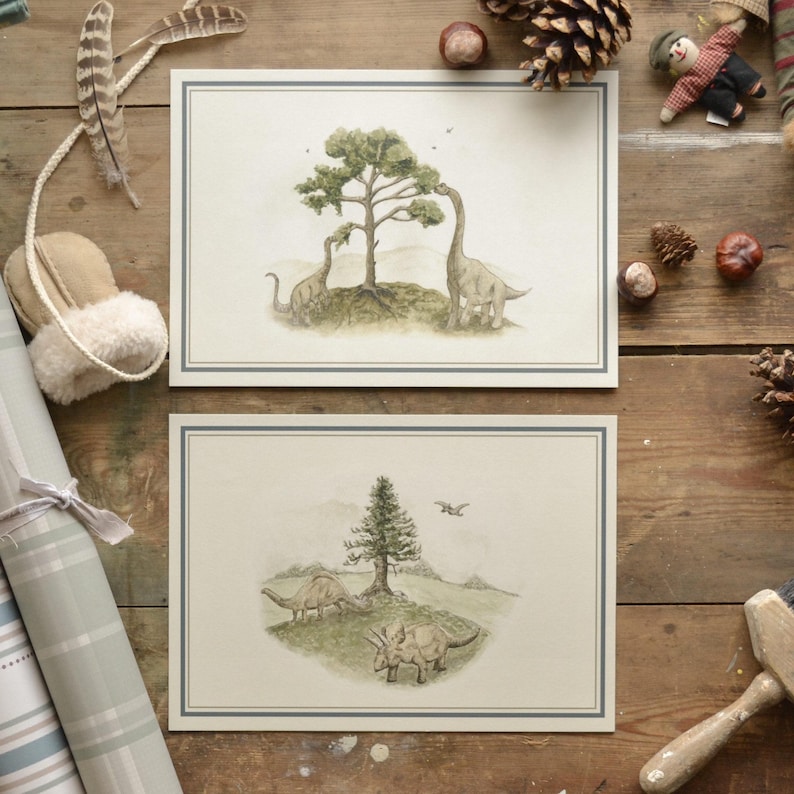 May include: Two dinosaur-themed art prints. The top print features two long-necked dinosaurs near a tree. The bottom print shows three dinosaurs near a tree. Both prints have a light blue border and are on a cream background. The prints are on a wooden surface.