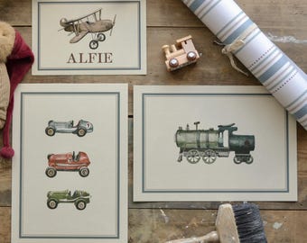 May include: Watercolor prints of vintage vehicles, including a biplane with the name "ALFIE", race cars, and a train. The prints are framed with a light blue border and set on a wooden surface. A wooden toy train and a paint brush are also visible.