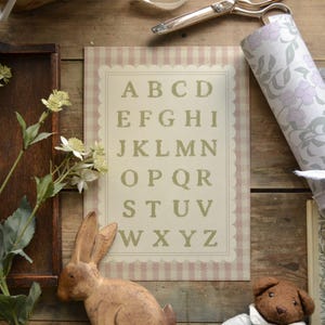 Op de afbeelding: Een decoratieve alfabetprint met een geschulpte rand en een roze en wit gestreepte achtergrond. De letters A t/m Z zijn gedrukt in een schreefletter. Een houten konijn en een teddybeer staan op de voorgrond, met bloemenpapier en een schaar op de achtergrond.