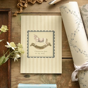 May include: A cream-colored card with vertical stripes features a framed illustration of a rocking horse. The illustration is in soft watercolor tones, with the name "Clementine Jane" and the date "10.05.2025" below. The card is on a wooden surface.
