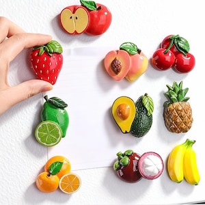 May include: A collection of colorful fruit-shaped refrigerator magnets. The magnets include a red apple, strawberry, peach, cherries, lime, avocado, pineapple, orange, mangosteen, and bananas. The magnets are attached to a white surface.