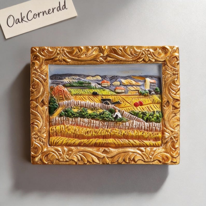 May include: A rectangular, gold-framed relief sculpture depicting a rural landscape with fields, houses, and figures. The frame has ornate, carved details. The text "OakCornerdd" is visible in the upper left corner.