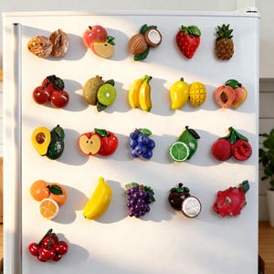 May include: A collection of colorful fruit-shaped refrigerator magnets. The magnets include a variety of fruits such as apples, bananas, strawberries, and grapes. The magnets are attached to a white refrigerator door.