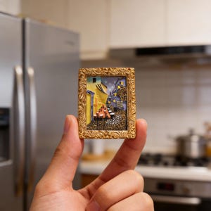 May include: A miniature framed artwork depicting a European street scene with cobblestone streets, buildings, and a starry night sky. The frame is gold-colored with ornate detailing. The artwork is held between fingers.
