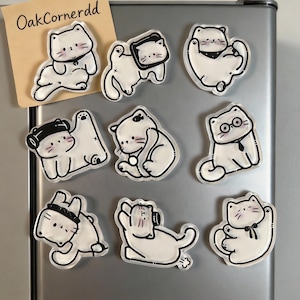 Puede incluir: Un conjunto de imanes transparentes con forma de gato, con contornos negros y varios accesorios. Los gatos están en diferentes poses, algunos con sombreros o gafas. También se ve un pequeño papel beige con el texto "OakCornerdd".