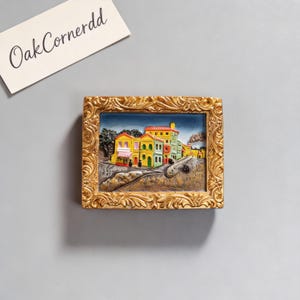 May include: A framed relief sculpture depicting a colorful village scene with yellow, green, and red buildings. The frame is ornate, gold-colored, and rectangular. The text "OakCornerdd" is on a white card in the upper left corner.