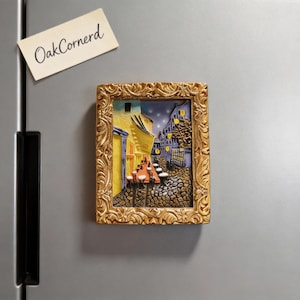 May include: A rectangular, gold-framed magnet featuring a 3D depiction of a European street scene at night. The artwork showcases buildings with yellow and blue hues, cobblestone streets, and tables with red tops. The magnet is attached to a gray surface, with a small note that reads "OakCornerd".