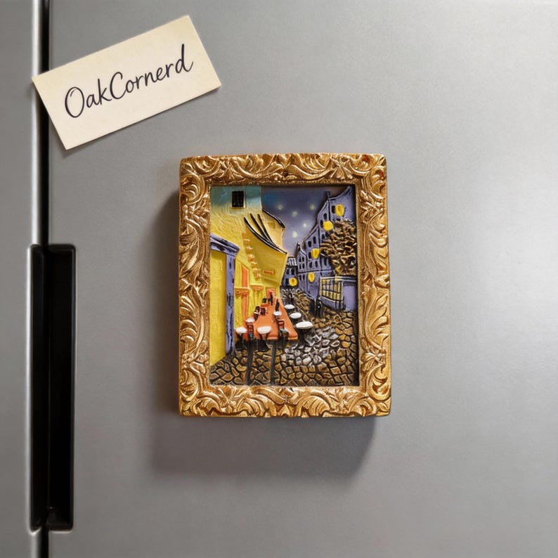 May include: A rectangular, gold-framed magnet featuring a 3D depiction of a European street scene at night. The artwork showcases buildings with yellow and blue hues, cobblestone streets, and tables with red tops. The magnet is attached to a gray surface, with a small note that reads "OakCornerd".