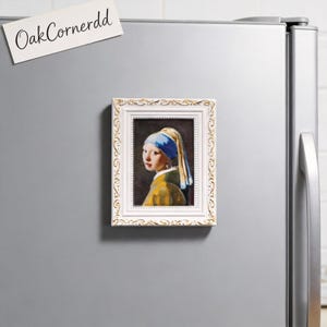May include: A framed magnet featuring a print of Johannes Vermeer's "Girl with a Pearl Earring." The white frame has a decorative gold pattern. The magnet is attached to a silver refrigerator. A white sticky note with "OakCornerdd" is attached to the refrigerator.