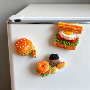 Op de afbeelding: Een verzameling kleurrijke koelkastmagneten met voedselthema's. Bevat een hamburger, een sandwich, een koffiekopje met de tekst "COFFEE" en andere miniatuurvoedselitems. De magneten zijn gedetailleerd en lijken van hars te zijn.