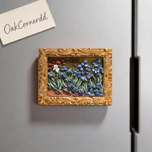 May include: A rectangular, gold-framed magnet featuring a bas-relief floral scene with blue and white flowers. The magnet is affixed to a silver refrigerator. A small note with the text "OakCornerdd" is also attached.