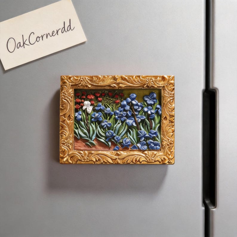 May include: A rectangular, gold-framed magnet featuring a bas-relief floral scene with blue and white flowers. The magnet is affixed to a silver refrigerator. A small note with the text "OakCornerdd" is also attached.