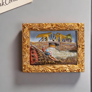 May include: A rectangular, gold-colored relief magnet depicting a bridge over a river with people in a boat. The bridge has yellow supports and a brick-like structure. The magnet is framed with an ornate gold border.