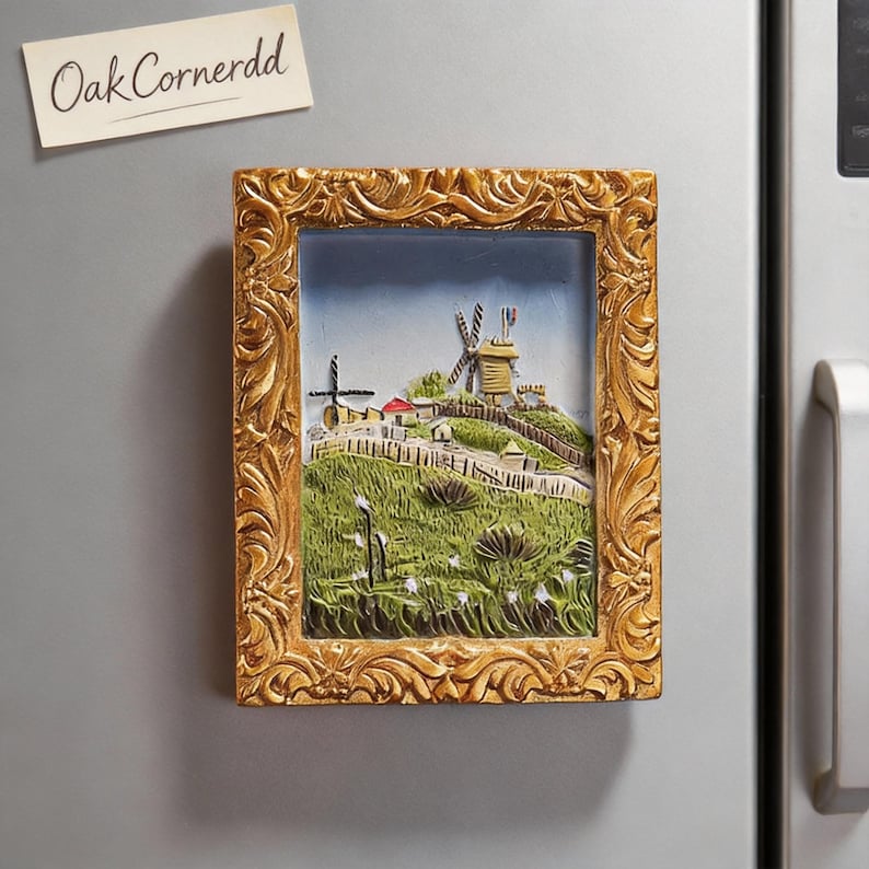 May include: A rectangular, gold-framed magnet featuring a 3D relief of a Dutch landscape with windmills, houses, and a grassy hill. The magnet is attached to a refrigerator. The text "Oak Cornerdd" is on a small, attached paper.