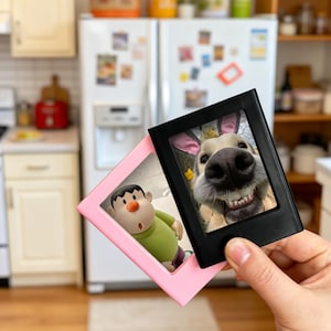 May include: Two small picture frames, one pink and one black, are held up in front of a kitchen. The pink frame contains a cartoon figure. The black frame displays a close-up photo of a dog wearing bunny ears. The dog is smiling.