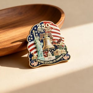 K&ouml;nnte beinhalten: Eine patriotische Anstecknadel mit der Freiheitsstatue, der Liberty Bell und dem Kapitol. Das Design beinhaltet die amerikanische Flagge und den Text "250th Anniversary 1776-2026" und "United States of America".