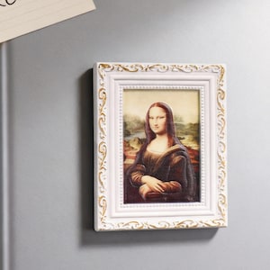 May include: A rectangular white framed magnet featuring a print of the Mona Lisa painting. The frame has a decorative gold-colored trim. The artwork depicts a woman with a subtle smile, set against a landscape background.