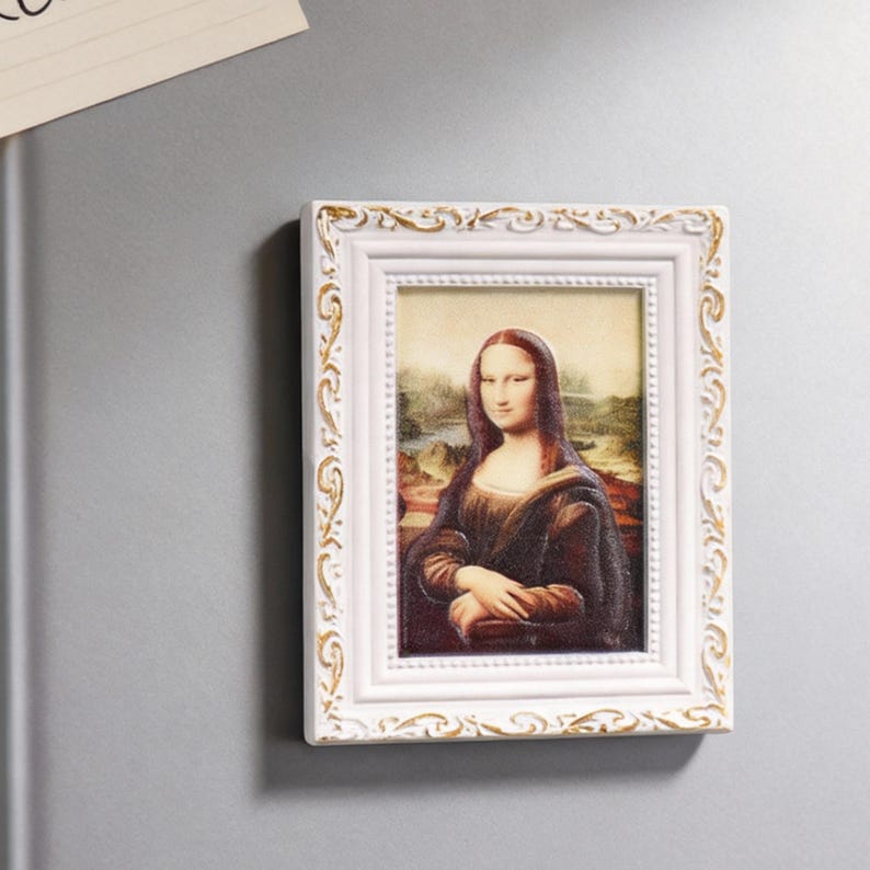 May include: A rectangular white framed magnet featuring a print of the Mona Lisa painting. The frame has a decorative gold-colored trim. The artwork depicts a woman with a subtle smile, set against a landscape background.