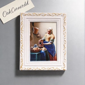 May include: A framed miniature of a painting depicting a woman pouring liquid from a jug. The frame is white with gold accents. The painting features a woman in a yellow and blue dress. The text "OakCornerdd" is on a white card.