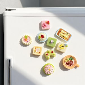 Könnte beinhalten: Eine Sammlung farbenfroher, miniaturisierter Kühlschrankmagnete zum Thema Essen. Die Magnete zeigen verschiedene Desserts und Gerichte, darunter Kuchen, Gebäck und eine Schüssel Reis. Die Magnete haben verschiedene Formen und Größen mit aufwendigen Details und leuchtenden Farben.
