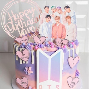 May include: A round, multi-layered cake with purple and pink frosting. The cake is decorated with a group of figures, heart-shaped toppers with names, and a glittery "Happy Birthday" cake topper. The cake has a BTS logo.