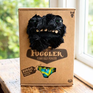 Op de afbeelding: Een bruine kartonnen doos met een zwart, harig monstergezicht met groene ogen. De doos heeft de tekst "Fugglers Funny Ugly Monster" en een label met "Budgie Fuggler Edition". Bevat een kleurrijke onderbroek.