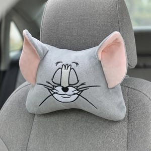 May include: Gray plush car headrest shaped like a cartoon mouse face. The headrest has large ears with pink inner ears, embroidered eyes, nose, and whiskers. It is designed to fit over a car seat headrest.