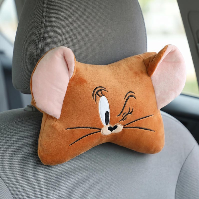 May include: A brown plush car headrest shaped like Jerry Mouse from Tom and Jerry. The headrest has large pink ears, a black and white eye, and a black nose and whiskers. It is attached to a gray car seat.