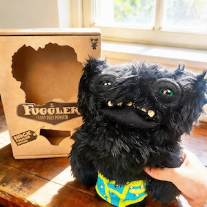 Könnte beinhalten: Ein schwarzes Fuggler-Plüschtier mit grünen Augen und einem grinsenden Mund, das blaue und gelb gemusterte Unterwäsche trägt. Das Spielzeug befindet sich neben seinem Karton, der das Fuggler-Logo und die Worte "Funny Ugly Monster" zeigt.