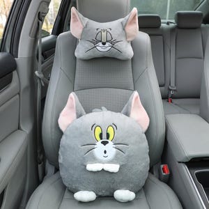 May include: Gray car seat headrest and cushion set featuring a cartoon cat face. The headrest is rectangular with large ears, and the cushion is round with a smiling cat face. The cat has yellow eyes, a white muzzle, and black whiskers.