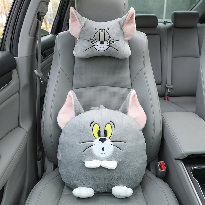 May include: Gray car seat headrest and cushion set featuring a cartoon cat face. The headrest is rectangular with large ears, and the cushion is round with a smiling cat face. The cat has yellow eyes, a white muzzle, and black whiskers.