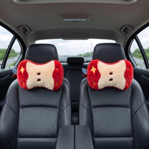 May include: Two car headrest pillows shaped like apples. The pillows are red with a beige center and have two brown button-like details. Each pillow has a yellow star and small green accents. The pillows are on black car seats.