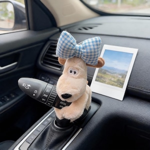 May include: A plush beige dog figurine with a black nose and white eyes featuring black pupils, wearing a blue and white gingham bow on its head. The plush toy is positioned around the turn signal lever of a car steering column.