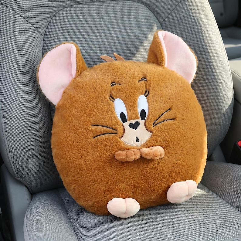 May include: A plush, round, brown Jerry the Mouse cushion with large pink-lined ears, white eyes, and a surprised expression. The cushion has small pink feet and is placed on a gray car seat.