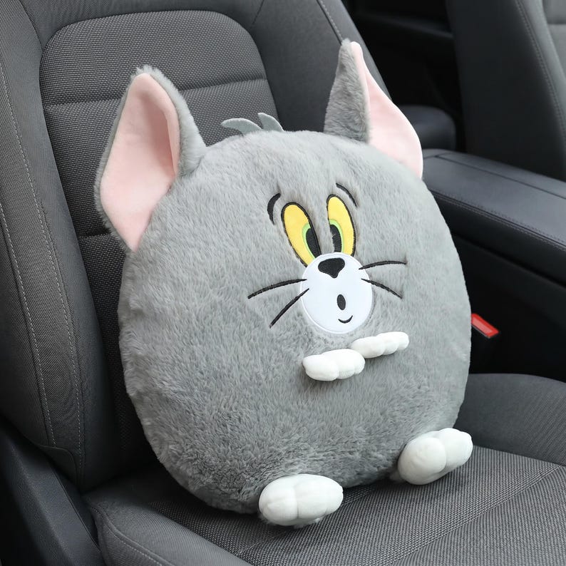 May include: A plush, gray cat-shaped pillow with pink inner ears, yellow eyes, and a white bow tie. The pillow is round and sits on a car seat, offering a whimsical design for car decor.