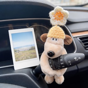 May include: A plush beige dog toy with brown ears and a black nose, wearing a yellow crocheted hat topped with a white and orange crocheted flower. The toy is wrapped around a black car turn signal lever next to a white-bordered instant photo.