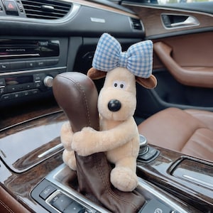May include: A plush dog toy with a blue and white checkered bow, brown ears, and a black nose, hugging a car's gear shift. The toy is beige and appears to be made of soft fabric. The car interior features brown leather seats and wood trim.