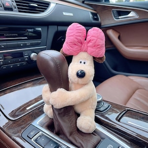 May include: A plush dog toy with a pink bow on its head, hugging a car gear shift. The dog has brown ears and a black nose. The interior of the car is visible, with brown leather seats and a wooden dashboard.