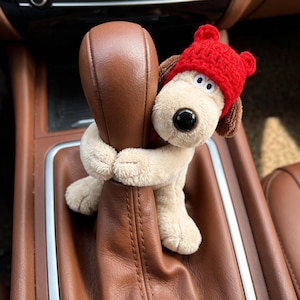 Op de afbeelding: Een pluchen hondenspeeltje met een rode gebreide muts die een bruine autoversnellingspook omhelst. De hond heeft een zwarte neus, witte ogen en bruine oren. Het auto-interieur is bruin.