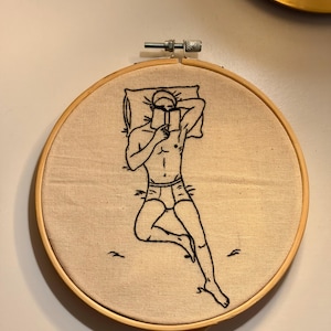 May include: Embroidered artwork in a wooden hoop. The design is a black line drawing of a person lying down, reading a book. The figure is wearing underwear and resting on a pillow. The background is beige fabric.
