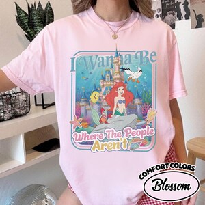 Puede incluir: Camiseta rosa claro con un gr&aacute;fico de Ariel de La Sirenita frente a un castillo. El texto "I Wanna Be Where The People Aren't" est&aacute; en un marco azul. La camiseta es Comfort Colors Blossom.