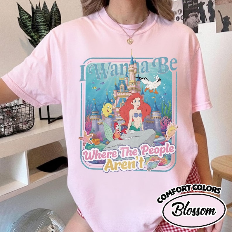 Puede incluir: Camiseta rosa claro con un gr&aacute;fico de Ariel de La Sirenita frente a un castillo. El texto "I Wanna Be Where The People Aren't" est&aacute; en un marco azul. La camiseta es Comfort Colors Blossom.