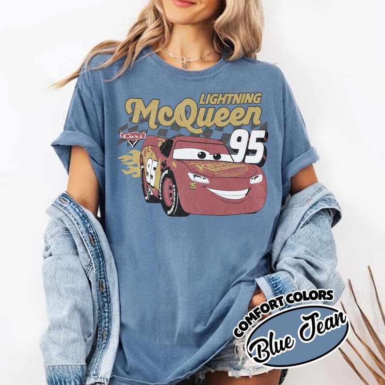 May include: Blue t-shirt featuring a graphic of Lightning McQueen from the movie Cars. The design includes the text "Lightning McQueen 95" in gold and white. The shirt is a comfort colors style.