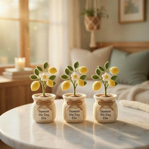 May include: Three crocheted lemon trees in small, beige pots. Each tree has yellow lemons, green leaves, and white flowers. The pots have a brown bow and a tag that reads "Squeeze the Day Ella". The scene is set on a white table.
