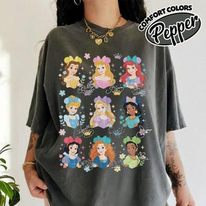 May include: Dark gray t-shirt featuring colorful illustrations of Disney princesses. The shirt has the words "COMFORT COLORS" and "Pepper" printed on the upper right corner. The princesses are Belle, Aurora, Ariel, Cinderella, Rapunzel, Jasmine, Snow White, and Merida.