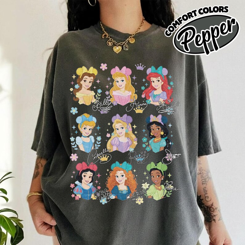 May include: Dark gray t-shirt featuring colorful illustrations of Disney princesses. The shirt has the words "COMFORT COLORS" and "Pepper" printed on the upper right corner. The princesses are Belle, Aurora, Ariel, Cinderella, Rapunzel, Jasmine, Snow White, and Merida.