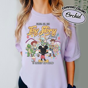 May include: Orchid-coloured t-shirt with a graphic of Toy Story characters and Mickey Mouse. The design includes the text "Toy Story Land" and "To Infinity and Beyond". The shirt is from Florida, established in 2018.