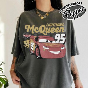 May include: Dark gray t-shirt featuring Lightning McQueen from the Cars movie. The design includes the text "Lightning McQueen 95" with a red race car graphic. The shirt also has the text "Comfort Colors Pepper" in a circular design.