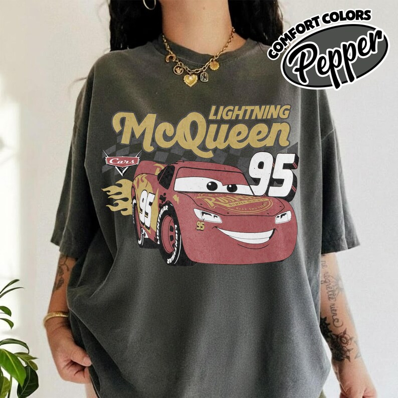 May include: Dark gray t-shirt featuring Lightning McQueen from the Cars movie. The design includes the text "Lightning McQueen 95" with a red race car graphic. The shirt also has the text "Comfort Colors Pepper" in a circular design.