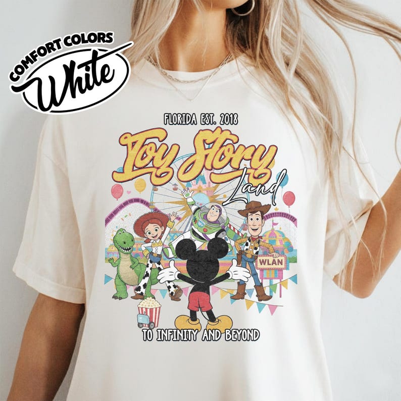 May include: Cream-coloured t-shirt with a vibrant graphic of Toy Story characters and Mickey Mouse. The design includes the text "Toy Story Land" and "To Infinity and Beyond". The shirt also has the text "Comfort Colors" and "Florida Est. 2018".